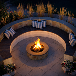Circular patio with lit fire pit, curved stone seating, cushions at night. | Down To Earth Landscaping | Jackson New Jersey, USA (732) 833-7702
