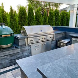 Stainless steel grill on a professionally installed patio and paver outdoor kitchen. Expansive modern house with porch and professionally installed and maintained lush landscaping, paved driveway, with plants and shrubbery near detached garage. | Down To Earth Landscaping | Jackson New Jersey, USA (732) 833-7702