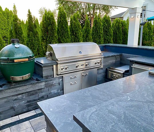 Stainless steel grill on a professionally installed patio and paver outdoor kitchen. Expansive modern house with porch and professionally installed and maintained lush landscaping, paved driveway, with plants and shrubbery near detached garage. | Down To Earth Landscaping | Jackson New Jersey, USA (732) 833-7702